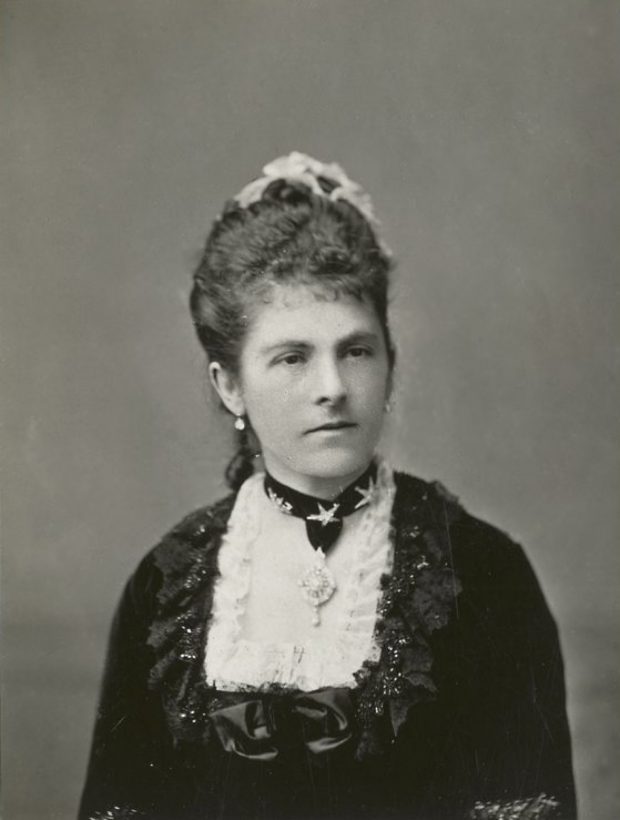 Photographie noir et blanc plan buste d'une femme (Lady Dufferin) dont les cheveux ont été coiffés; la dame est vêtue d'une robe de cérémonie à corsage en dentelle carré, parée d’un collier-ruban orné d’un bijou. 