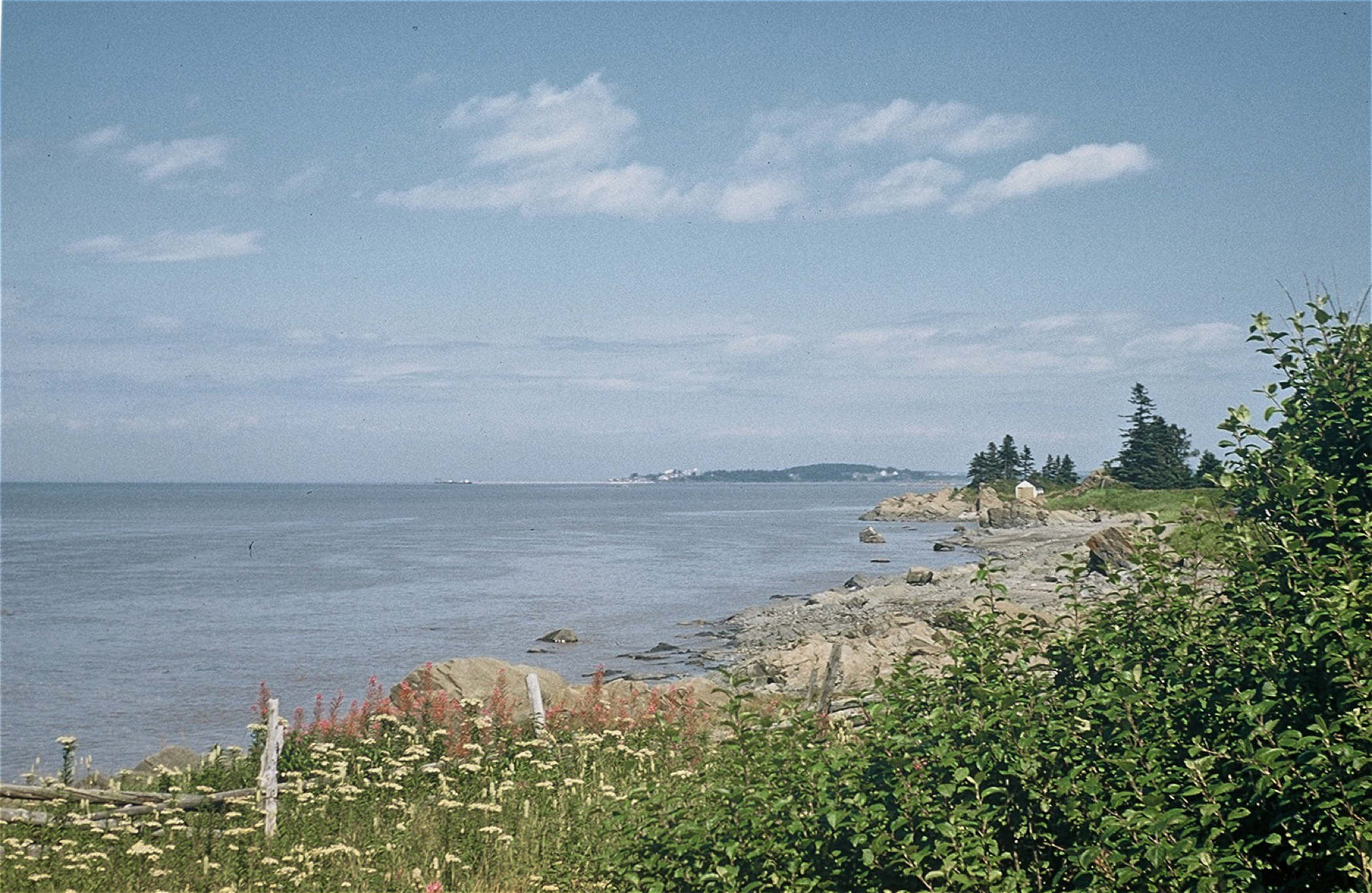 Photographie couleur d’un paysage fluvial (Rivière-du-Loup) où l’on aperçoit au premier plan une clôture en bois, des rochers, un rivage sablonneux et des arbustes en bordure du fleuve St-Laurent s’étendant sous un ciel bleu nuageux.