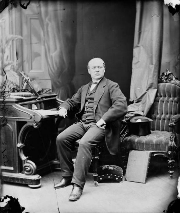 Photo noir et blanc d'un homme d'âge moyen (Sir Thomas McGreevy), partiellement chauve, assis sur une chaise capitonnée, son bras droit appuyé sur un secrétaire; son regard tourné vers la caméra. Il est vêtu d’un costume officiel sombre de style mi-victorien; un chapeau haut-de-forme repose à l'envers sur une autre chaise en velours capitonnée à ses côtés. En arrière-plan, des tentures suspendues dont une devant une porte. 