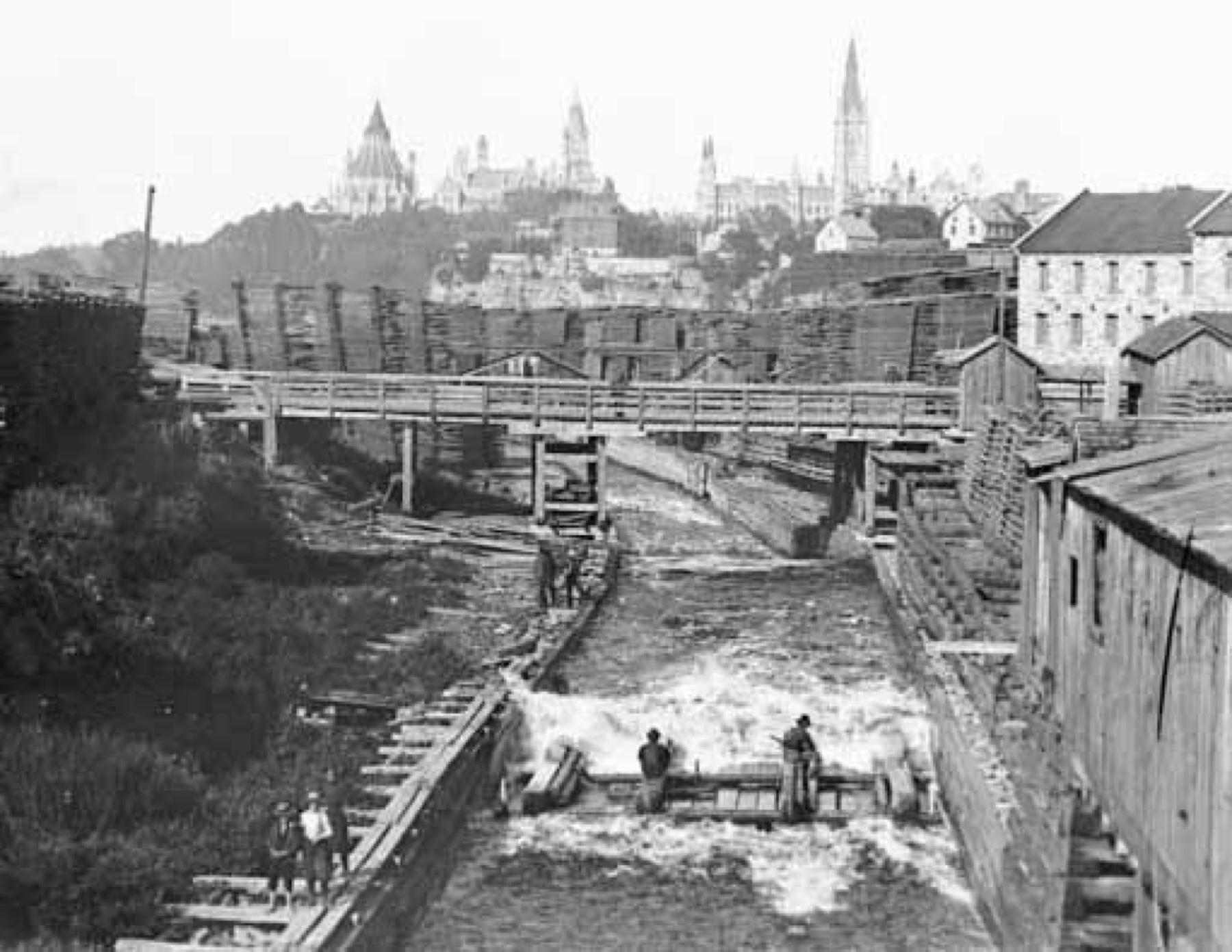 Photographie noir et blanc d’une scierie au-dessus d'un canal avec des bâtiments du Parlement d’Ottawa en arrière-plan; dans le canal, des hommes à l’œuvre sur un radeau de bûcheron.