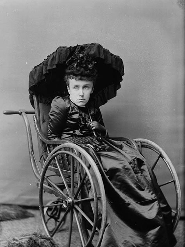 Photo noir et blanc d'une femme adulte (Mary Macdonald) affichant une expression triste et inhabitée, assise dans un fauteuil roulant du XIXe siècle et tenant un parasol noir ouvert au-dessus de sa tête. 