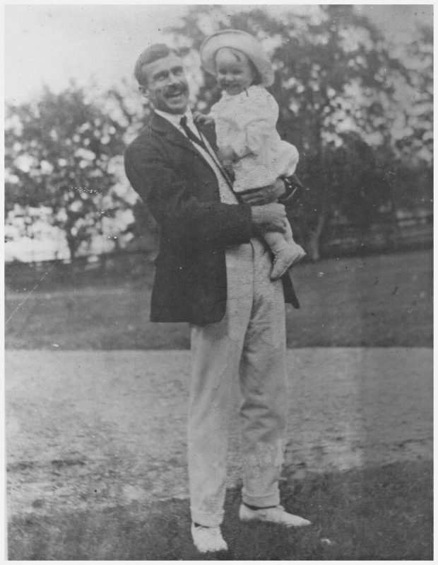 Photographie noir et blanc d'un homme de grande taille (Kenneth Molson), souriant, tenant dans ses bras un enfant en bas âge, lui aussi souriant, posant à l'extérieur sur une pelouse devant une rangée d’arbres en arrière-plan. 
