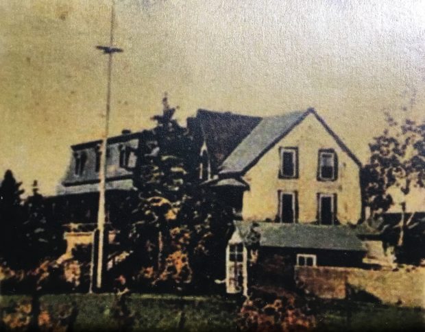 Photographie colorée à la main (à l'origine en noir et blanc) d'une grande maison (la villa Les Rochers) devant laquelle un mât de drapeau est érigé. Sur le côté de la propriété, on aperçoit une remise.