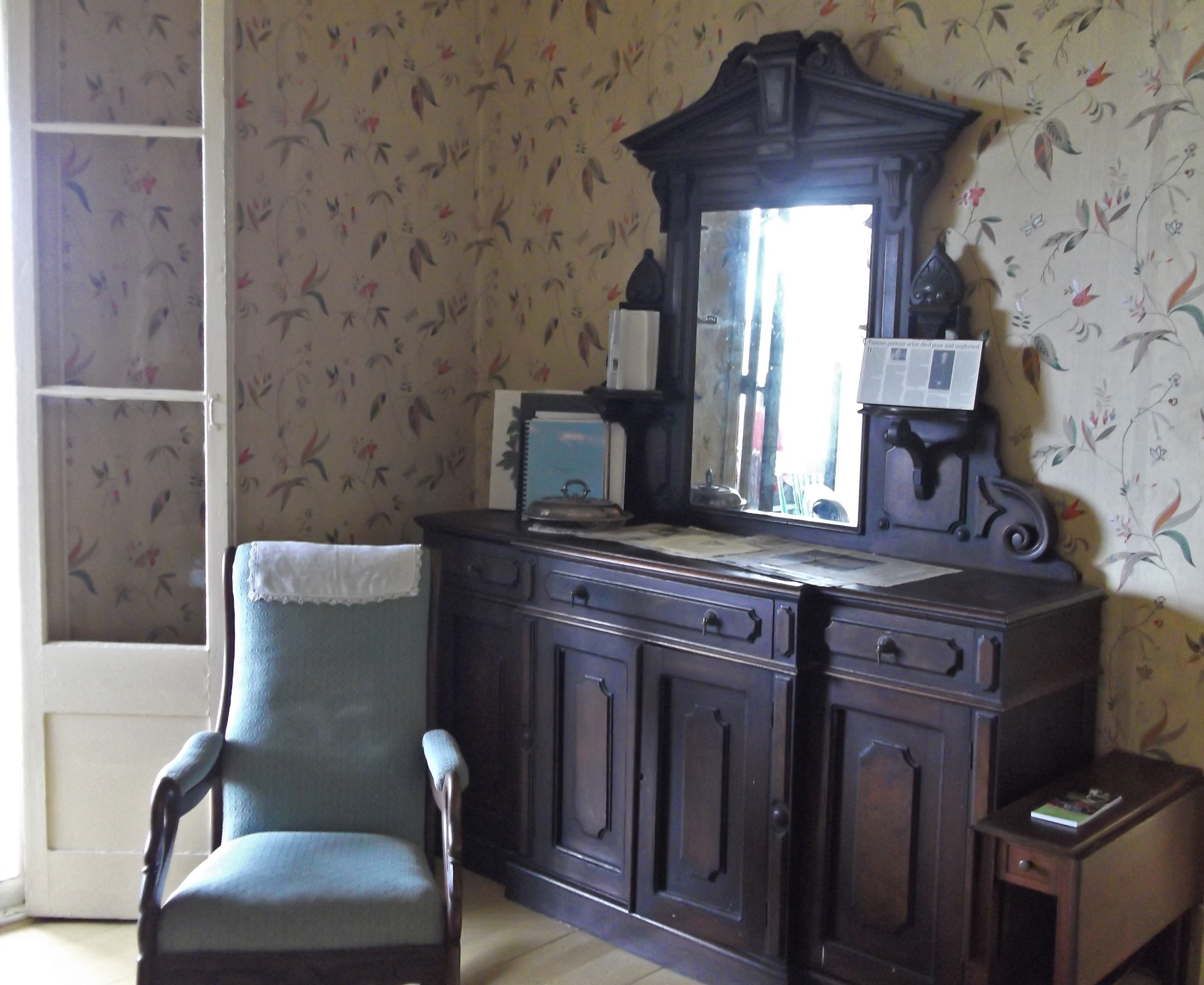 Vue intérieure d’un coin de salle à manger meublée d’une chaise capitonnée et d’un buffet en bois avec tiroirs et portes, surmonté d’un miroir, adossé à un mur recouvert de papier peint à motifs floraux. 