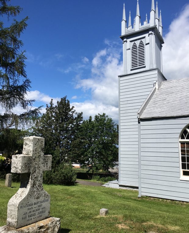 Photographie couleur de l'église St. Bartholomew, une vieille église en bois avec un clocher en façade vue de côté. Sur la photo apparait au premier plan une pierre tombale et à l'arrière-plan, des arbres sous un ciel bleu.