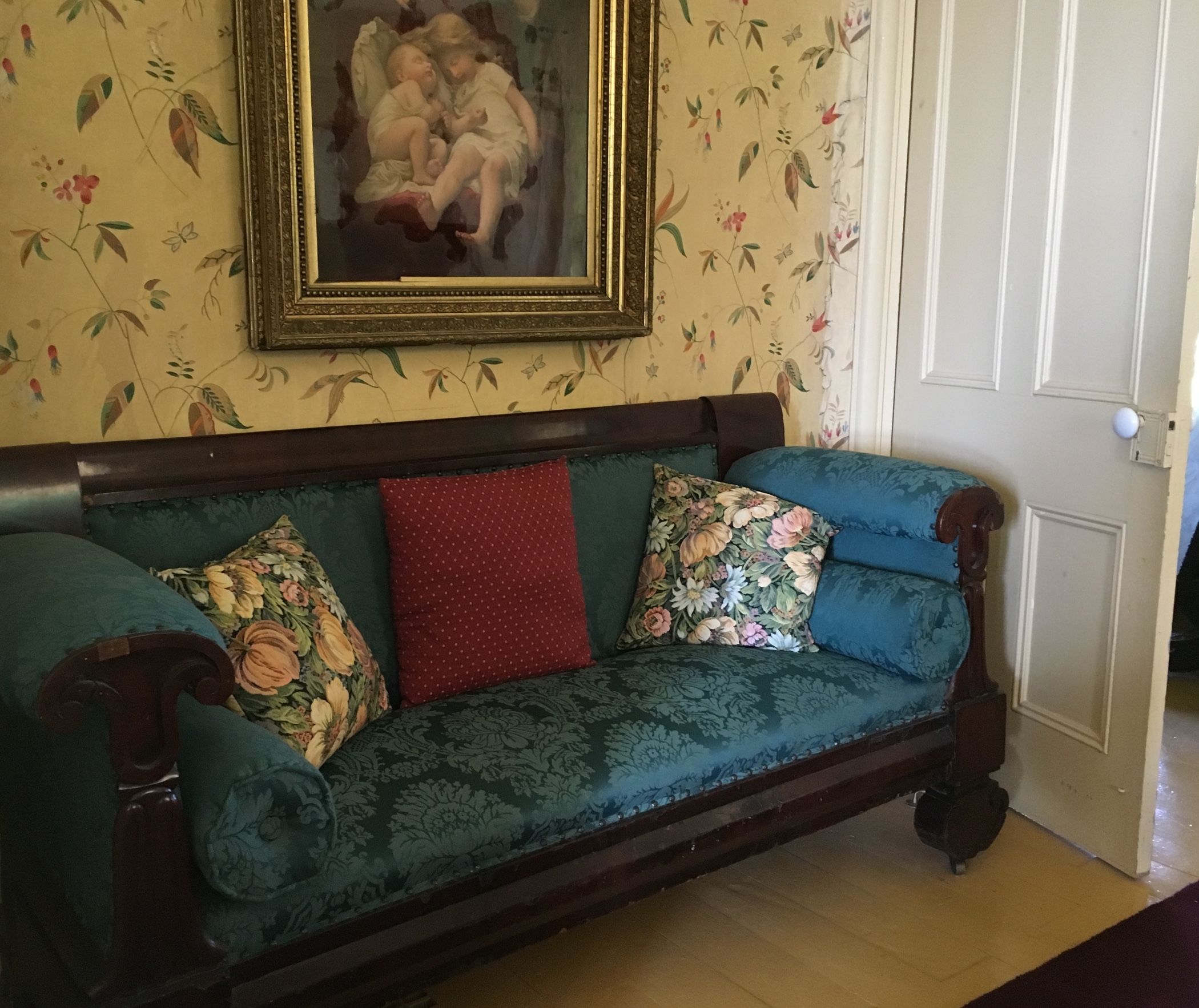Une photo couleur d'un vieux sofa Chesterfield capitonné installé dans le coin d’une pièce derrière une porte, le dos contre un mur recouvert de papier peint sur lequel est accroché un tableau illustrant des chérubins. 