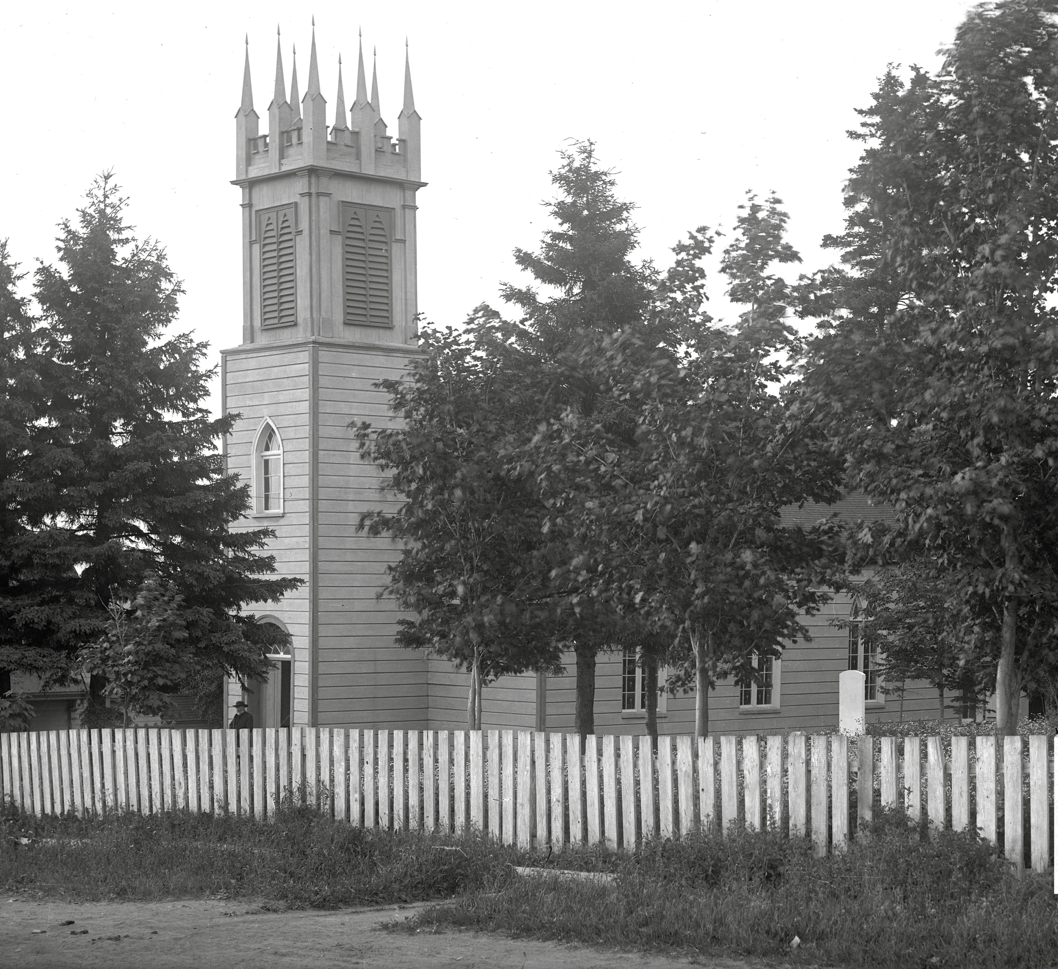Photo noir et blanc d'une vieille église en bois, église anglicane St. Bartholomew, surmontée d’un clocher en façade; un prêtre sortant de son entrée principale. Au premier plan, une clôture en bois sur le fronton de l'église, derrière laquelle se dressent de grands arbres.