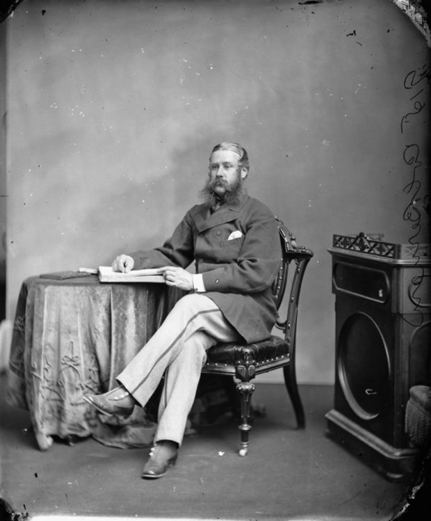 Portrait noir et blanc de Sir Hewitt Bernard assis, vêtu d'un manteau court en laine et d'un pantalon à rayures, les jambes croisées, son bras droit appuyé sur un livre ouvert déposé sur une table ronde. 