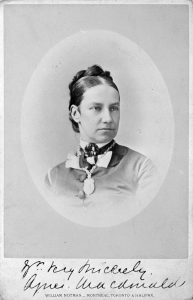 A head-and-shoulders portrait of a 30-35-year-old woman, her dark hair pulled tightly into an arrangement of braids pinned behind her head, She wears a buttoned-up shirt, a cameo at her throat and a necklace the details of which are indistinguishable. Her expression is a mixture of thoughtful and stern.