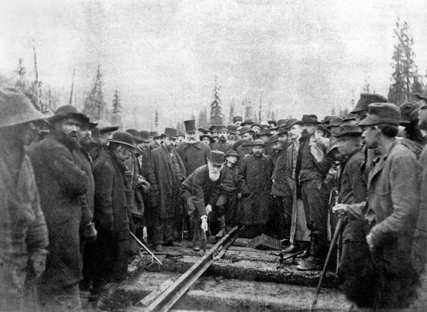 Une photo en noir et blanc datant de 1885, montrant plus d'une centaine d’hommes revêtus de manteaux de laine entourant un homme, Sir Donald A. Smith, coiffé d'un haut-de-forme, penché au-dessus d’une voie ferrée, muni d’un pic, enfonçant le dernier crampon du chemin de fer Canadien Pacifique.