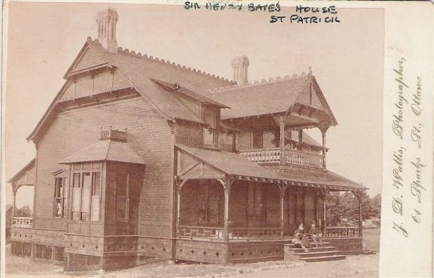 Vieille carte postale montrant une maison antique en bois à deux étages parée de boiseries et garnitures finement ciselées; deux personnes étant assises sur les marches de sa véranda. 