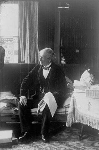 Sir John A. Macdonald en costume officiel, assis sur un canapé, tenant une liasse de papiers déposés sur son genou. 