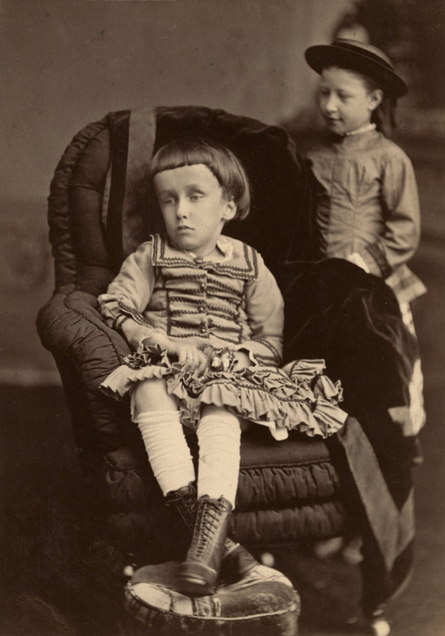 Photographie noir et blanc d'une petite fille (la jeune Mary Macdonald), assise sur une chaise, les jambes croisées devant elle, l'air absent. On dénote que sa tête est anormalement grosse, auréolée d’une chevelure noire coupée au carré avec une frange sur son large front. 
