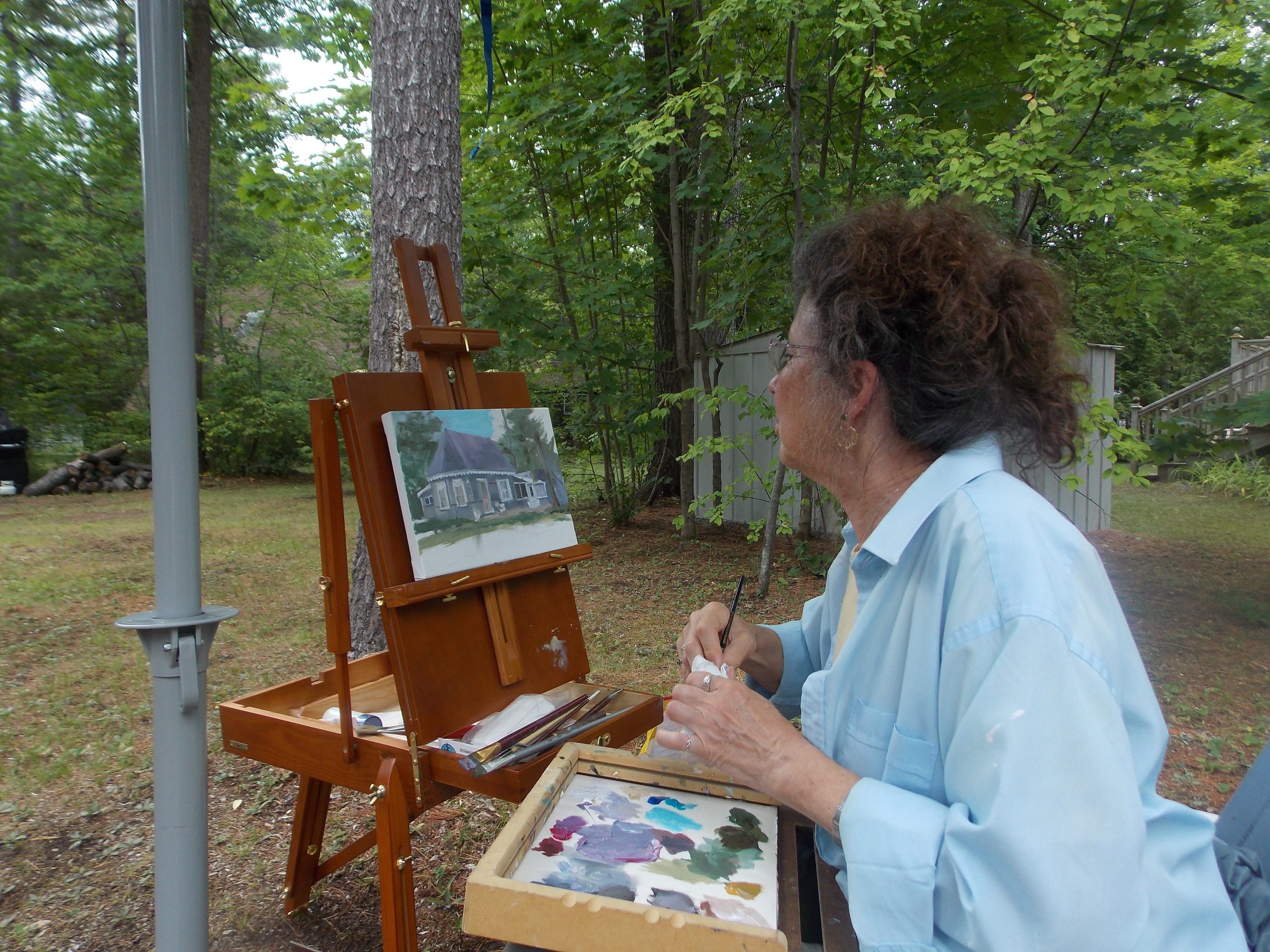 Image de quelqu’un assis dehors devant un chevalet, qui y peint un chalet. 