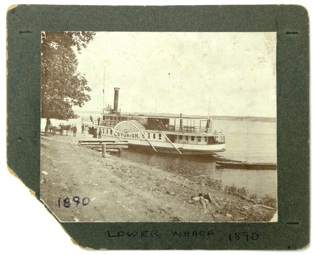 Image d’un grand bateau à vapeur à côté du quai, avec des gens et un cheval et chariot, sur le quai entre un grand arbre et le bateau.