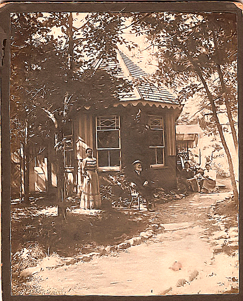 Image d’un homme portant un chapeau de paille, assis les jambes croisées devant un chalet entouré de grands arbres, avec une femme debout à sa droite et d’autres personnes assises à l’arrière-plan. 