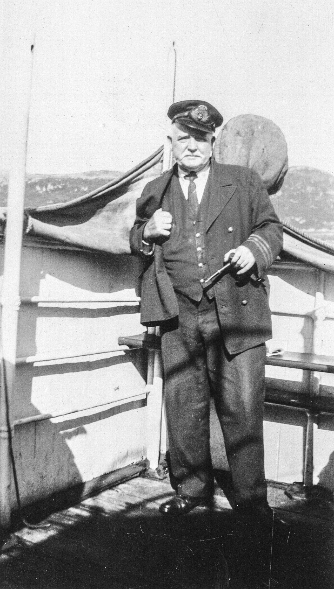 Photographie d'archives en noir et blanc d'un homme en uniforme avec une casquette à visière debout sur le pont d'un bateau tenant une pipe dans sa main gauche. Il y a des collines en arrière-plan.