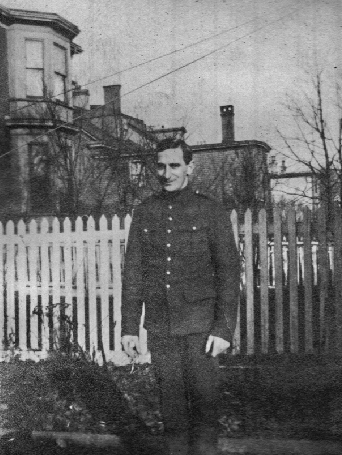Photographie d’archive en noir et blanc d’un homme en uniforme debout devant une clôture en piquets blancs située devant des maisons, des arbres et des fils électriques.
