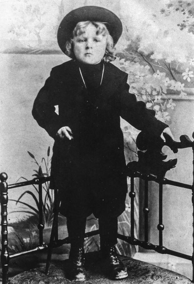Photographie d’archive en noir et blanc d’un garçon en habit sombre et chapeau à larges bords debout devant une balustrade dans un studio de photographe. 