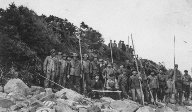 Photographie d’archive en noir et blanc. Un groupe de vingt hommes en habits et chapeaux de pluie, en combinaison ou en complet-cravate, tenant de longues perches sur une plage. Sur la colline derrière le groupe d’hommes se trouvent deux autres groupes d’hommes : à gauche, quatre hommes sont assis sur la colline et à droite, sept hommes se tiennent debout.