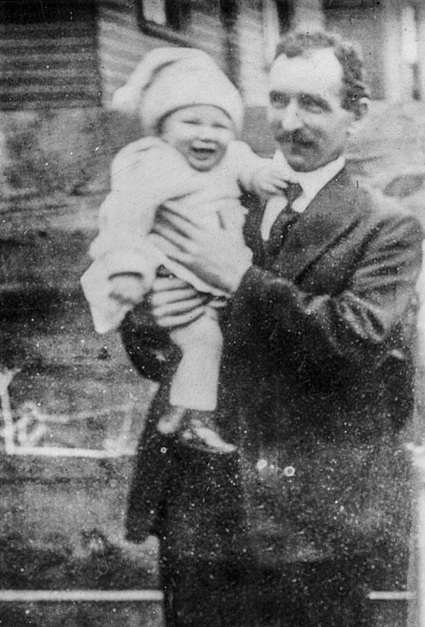 Photographie d’archive en noir et blanc d’un homme en costume-cravate debout à l’extérieur d’un bâtiment et tenant un bébé vêtu d’un chandail léger, d’un pantalon et d’un chapeau.