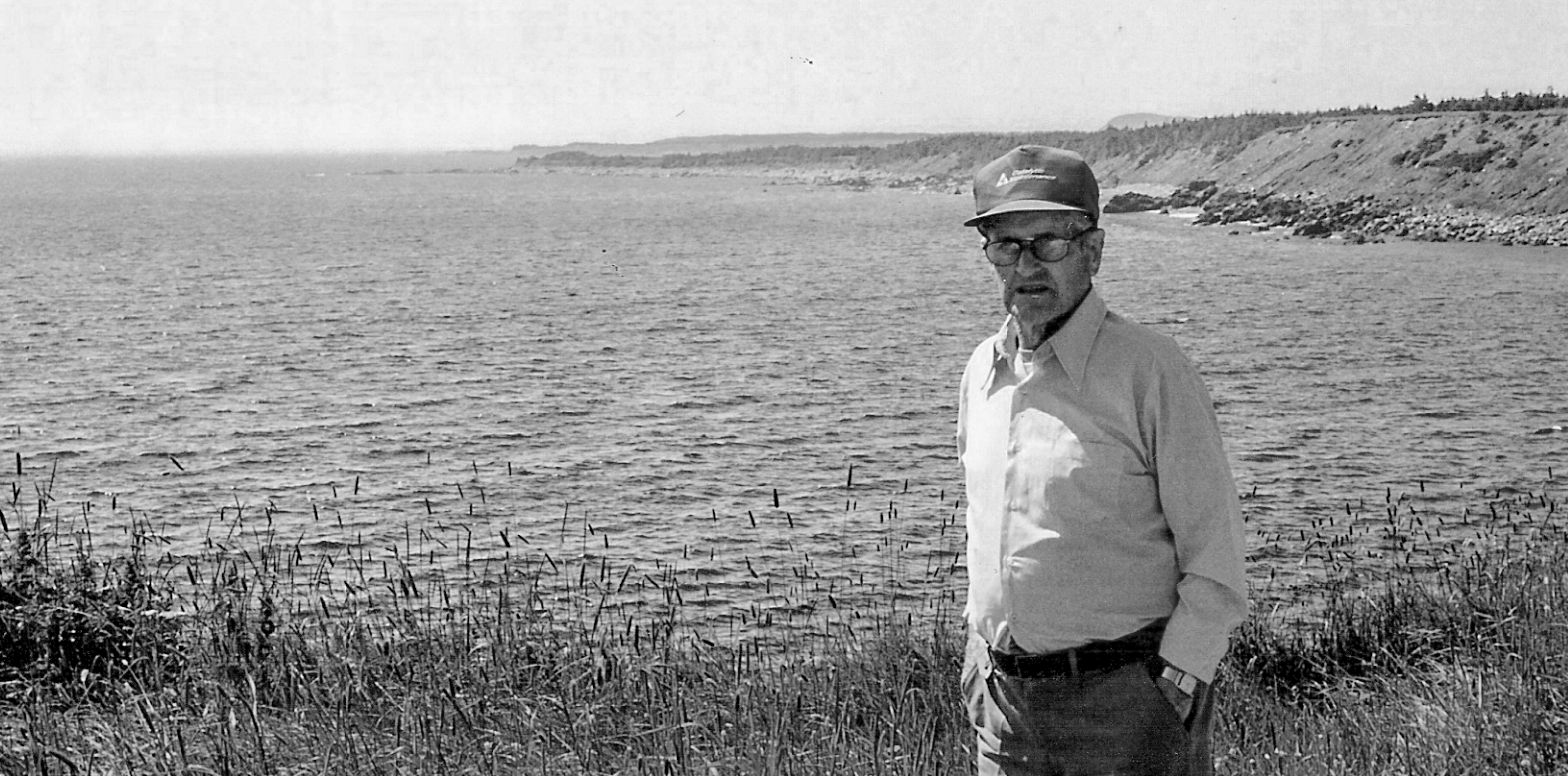 Photographie en noir et blanc d’un homme d’âge mûr portant des lunettes, une casquette de baseball, une chemise claire et des pantalons foncés. L’homme se tient dans un pré devant un paysage d’océan avec des collines à l’arrière-plan à droite.
