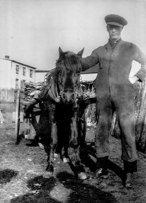 Photographie en noir et blanc d’un homme d’âge mûr portant des lunettes, une casquette de baseball, une chemise claire et des pantalons foncés. L’homme se tient dans un pré devant un paysage d’océan avec des collines à l’arrière-plan à droite.