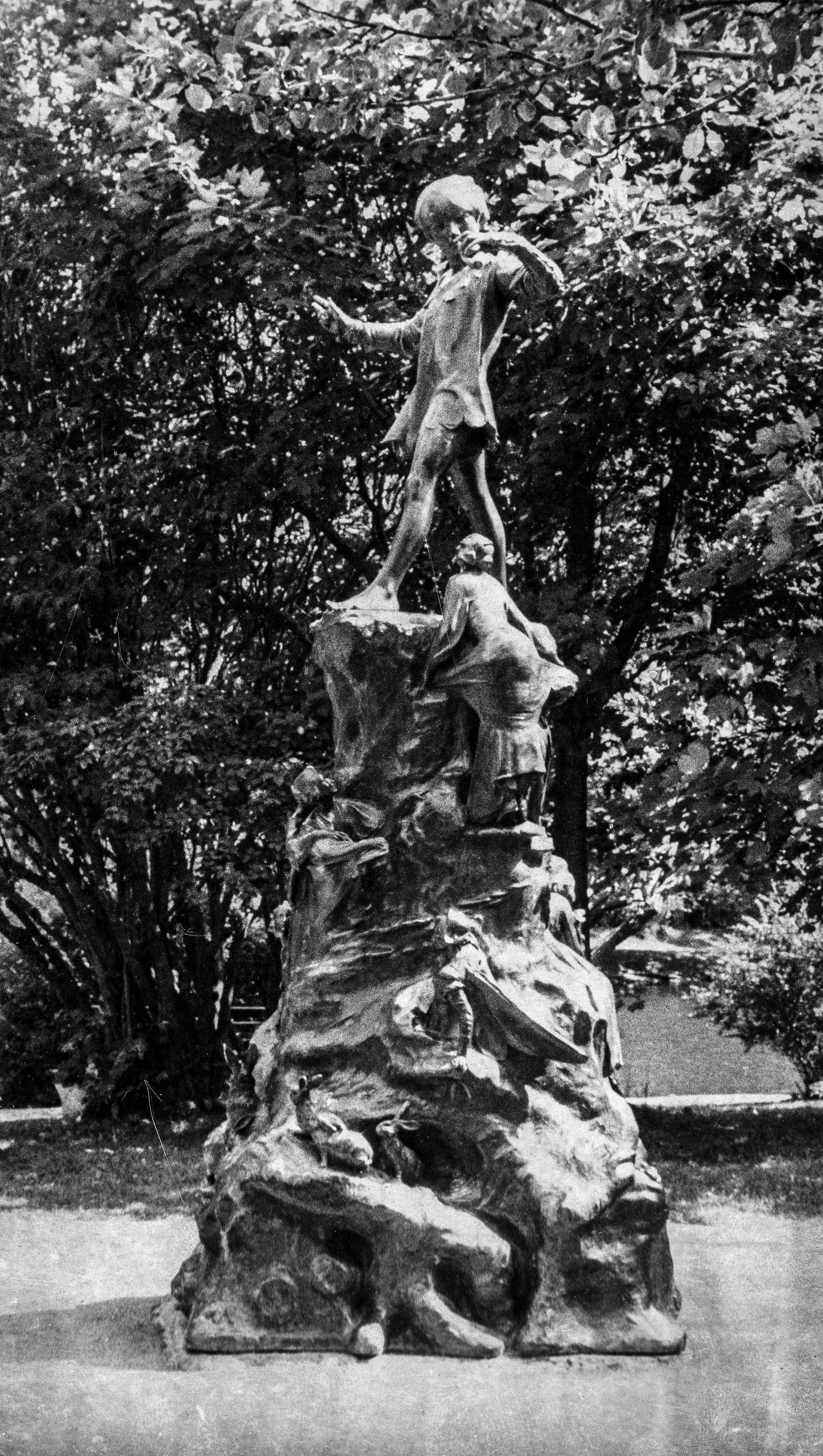 Photographie en noir et blanc de la statue de Peter Pan. Grande statue avec des animaux, des gens et un garçon se tenant debout au sommet.
