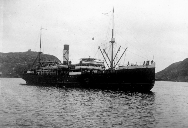Photographie d’archive en noir et blanc. Le grand paquebot SS Florizel dans le port de St. John’s. Derrière le bateau à gauche, on voit une colline avec une tour en pierre, et à droite des collines. Des gens sont visibles sur le pont du navire.