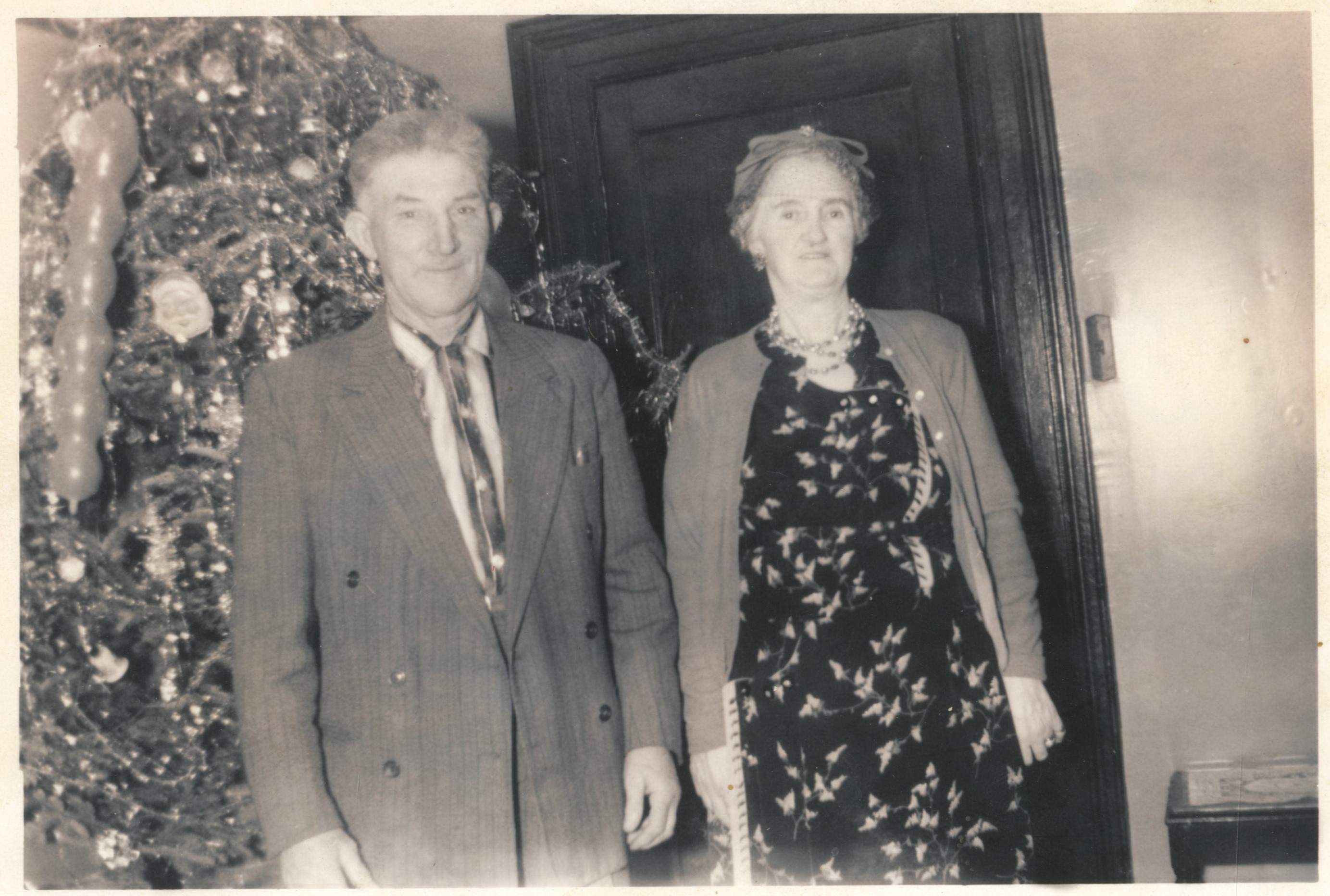 Photographie d’archive en noir et blanc d’un homme en costume et d’une femme portant une robe, un cardigan, un chapeau et un collier devant un arbre de Noël couvert de guirlandes et de décorations.