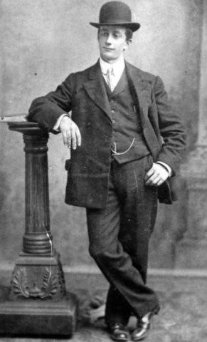 Photographie d’archive en noir et blanc d’un homme en costume-cravate et chapeau melon appuyé contre un poteau.