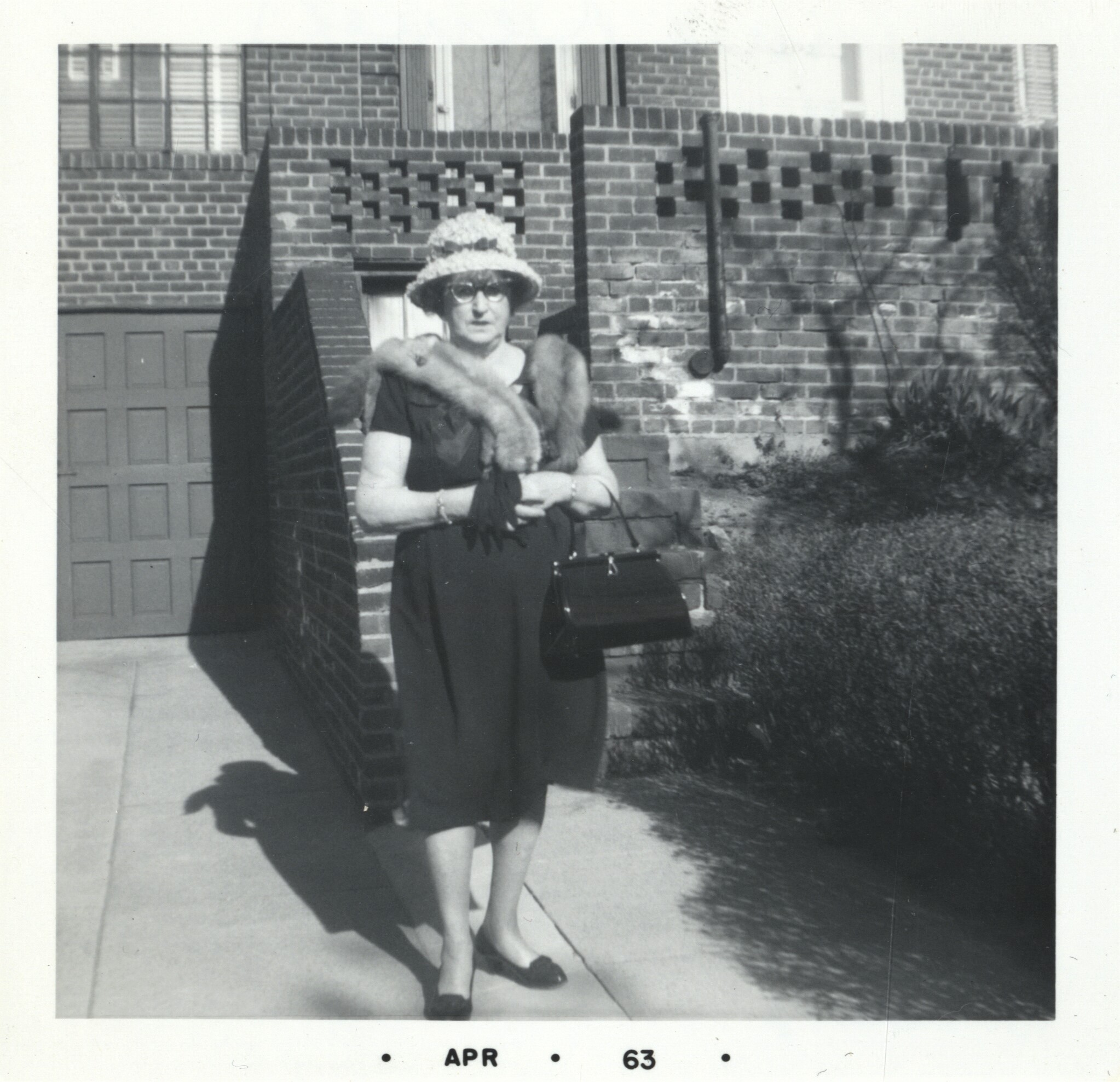 Photographie d’archive en noir et blanc d’une femme debout à l’extérieur d’un bâtiment en brique, portant une robe, un col de fourrure, un chapeau et des lunettes et tenant des gants et un sac à main.