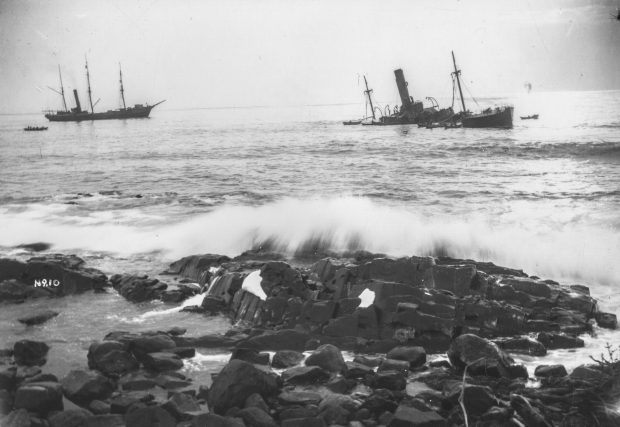 Photographie d’archive en noir et blanc d’un grand paquebot, le SS Florizel, échoué dans l’océan juste au large d’une plage rocheuse. On peut voir sept bateaux de sauvetage autour du Florizel. Un grand bateau de sauvetage peut être observé à gauche du Florizel, accompagné de deux plus petits bateaux de secours sur sa gauche.