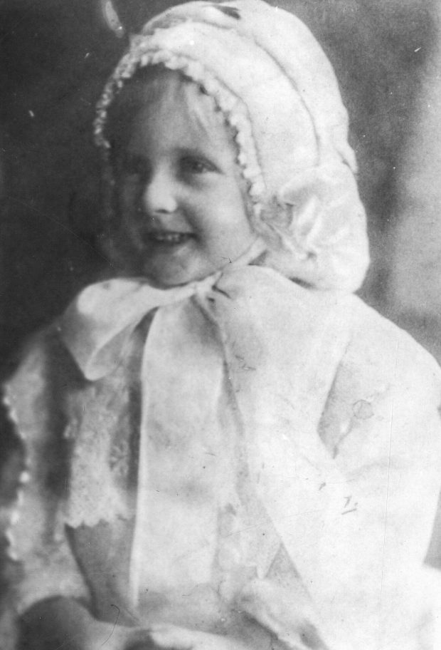 Photographie d’archive en noir et blanc d’une fille portant un bonnet blanc attaché par un nœud sous son menton ainsi qu’une robe blanche.