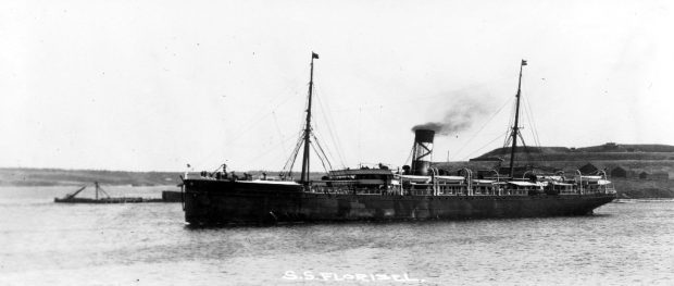 Photographie d’archive en noir et blanc. Le grand paquebot SS Florizel dans un port. Derrière le bateau à droite, on voit des maisons sur une colline, et à gauche un quai.