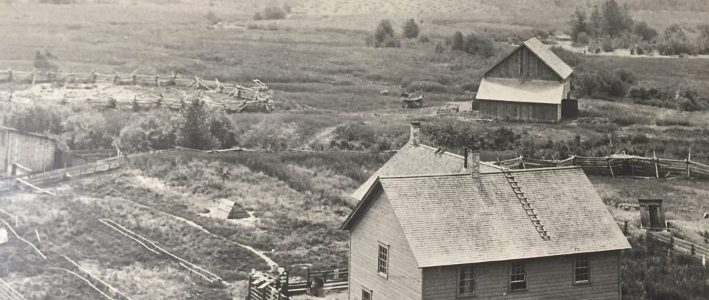 Une maison de deux étages est au centre d’une propriété pionnière. La propriété est entourée par des herbes du marécage et des arbustes. La maison est entourée par une clôture en bois. Il y a une étable et des corrals dans l’arrière-plan. Au-delà de l’étable, il y a un champ de foin.