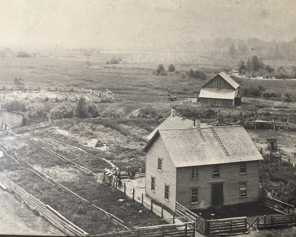 Une maison de deux étages est au centre d’une propriété pionnière. La propriété est entourée par des herbes du marécage et des arbustes. La maison est entourée par une clôture en bois. Il y a une étable et des corrals dans l’arrière-plan. Au-delà de l’étable, il y a un champ de foin.