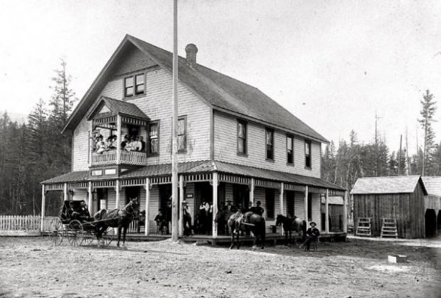 Un buggy et des chevaux sont devant un bâtiment en bois. Dans la deuxième histoire, quelques personnes sont sur un balcon. Plusieurs personnes sont rassemblées sur le balcon recouvert. Un homme est assis sur une chaise sur le chemin de terre.