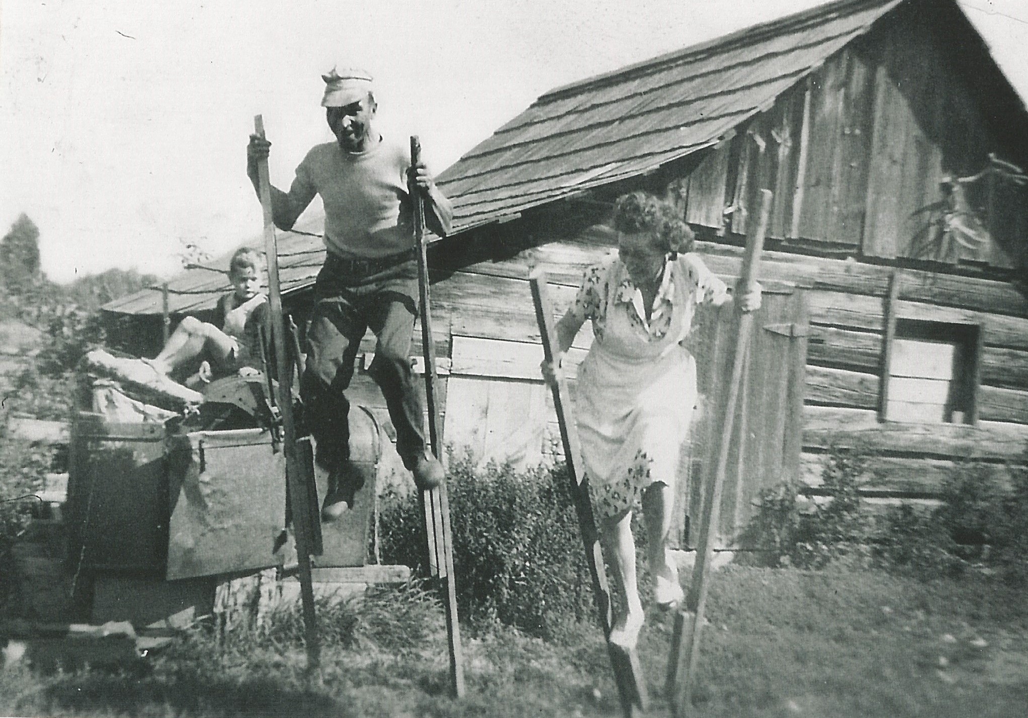 Un homme et une femme en robe jouent avec des pilotis en bois. Ils rient. Un garçon les regarde dans l’arrière-plan. Il y a une construction en dans l’arrière-plan qui semble être une étable.