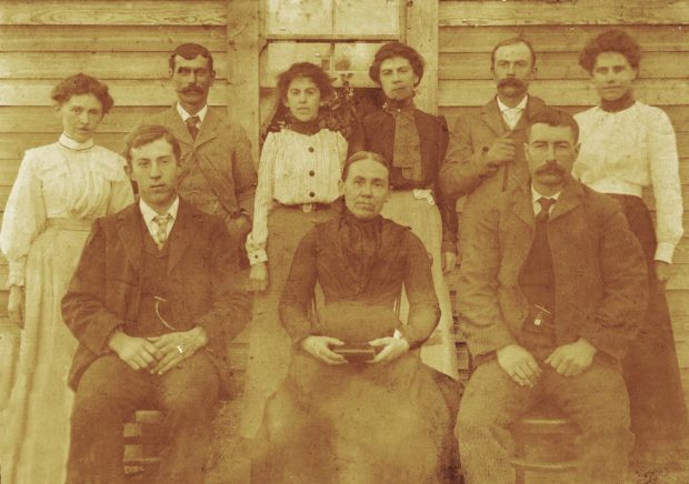 Un groupe de pionniers posent devant une construction en bois. Trois personnes sont assises sur des chaises, et le reste se tient debout. Une vieille dame est assise à la chaise au milieu et elle tient un livre.