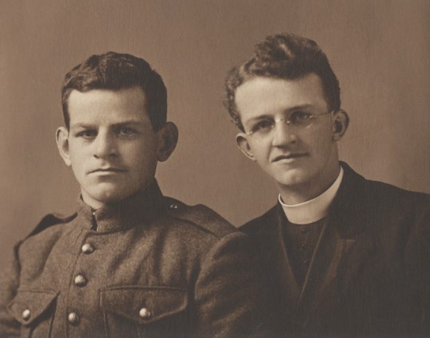 Deux jeunes hommes sont assis. Ils semblent être frères. Celui à la gauche a du cheveu foncé et a l’air sérieux. Il porte un uniforme de la Première Guerre mondiale. Celui à la droite est prêtre. Il porte des grosses lunettes en format de cercle. Ces cheveux sont ondulés. Il porte un col romain et des vêtements noirs.