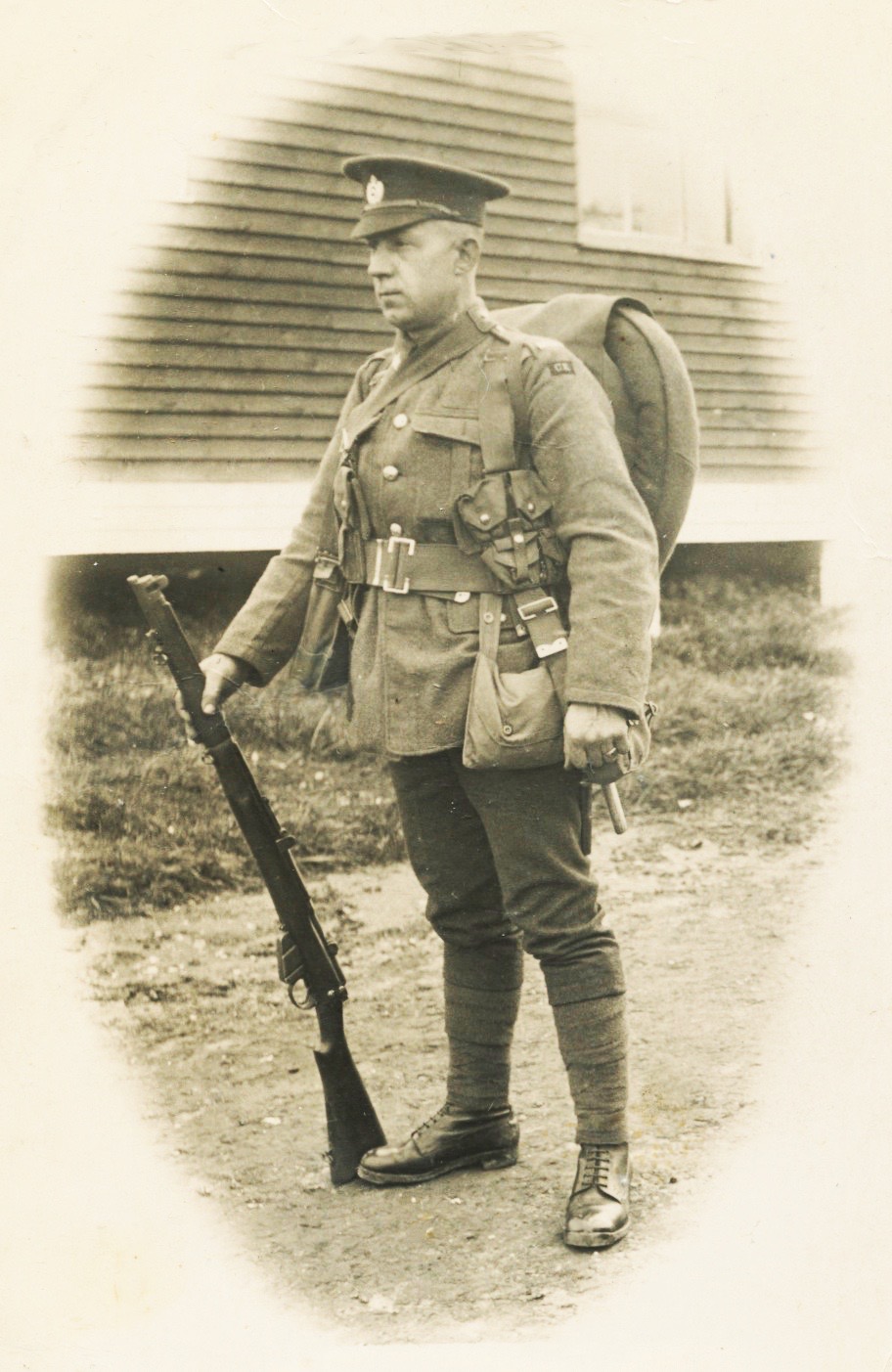 Un soldat est debout sur un chemin de terre. Il a un gros sac à dos. Il tient un fusil. 