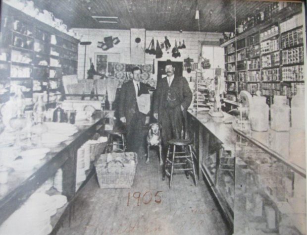 Deux hommes et un chien sont dans un magasin pionnier. Les murs sont couverts par des étagères. Les étagères, les comptoirs et le plancher sont couverts de marchandises. 