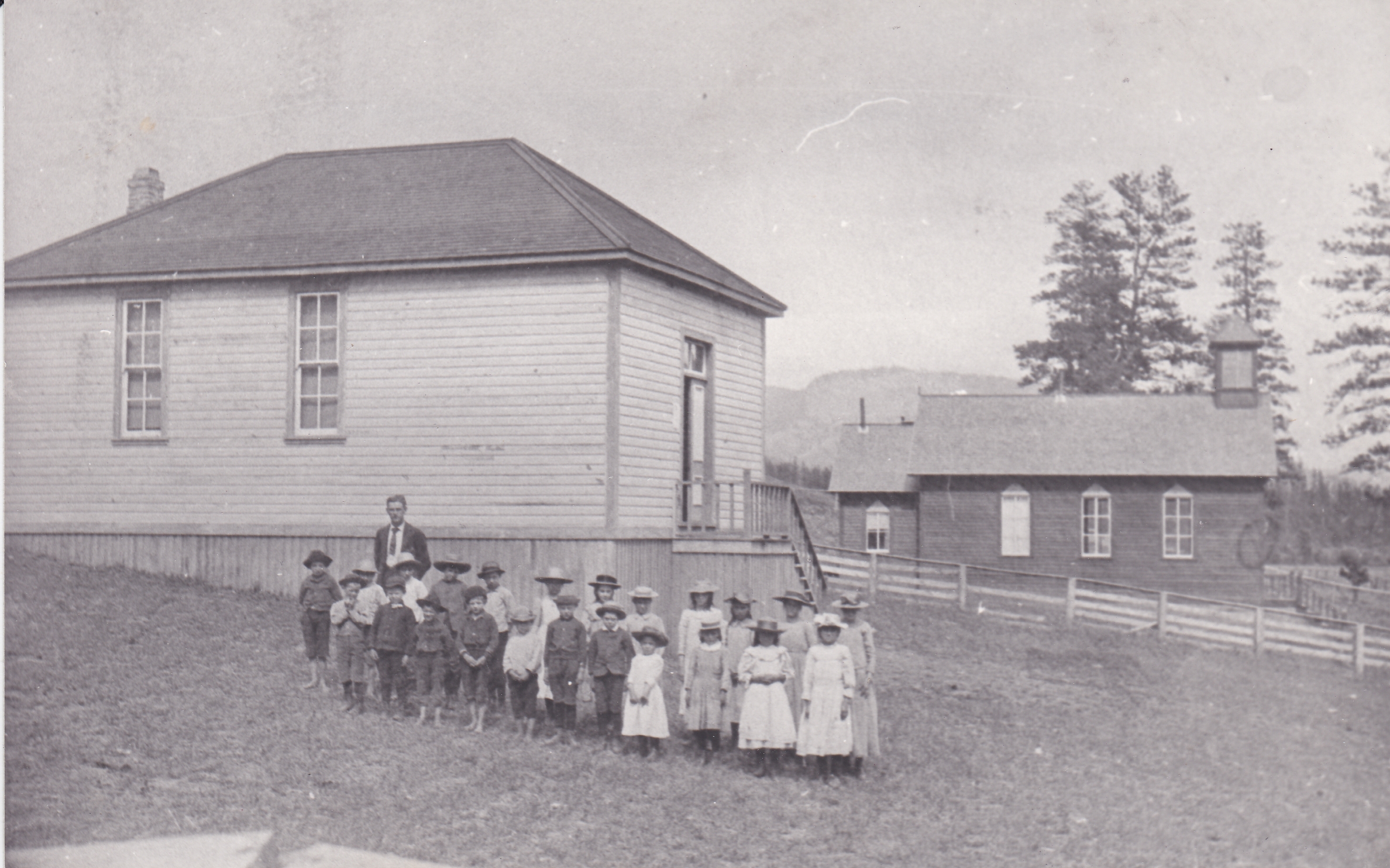 Un groupe d’enfants pionniers et leur professeur sont debout devant une école en bois. Tous les enfants portent des chapeaux. Il y a une église en bois à côté de l’école. 