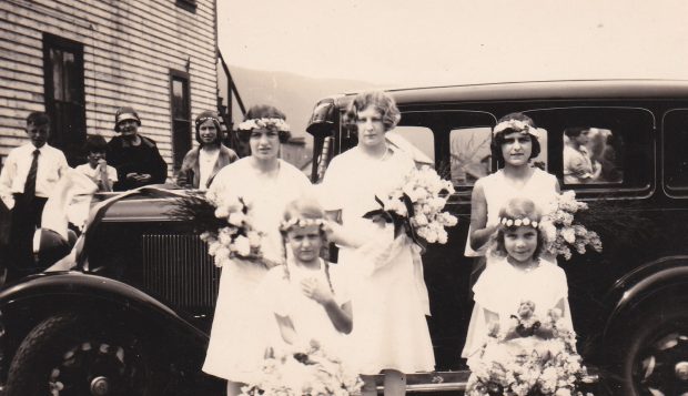 Cinq filles qui portent des robes blanches sont debout à côté d’une vieille voiture. La plupart ont des couronnes de fleurs dans les cheveux et elles tiennent des bouquets de fleurs. Deux des filles sont jeunes et on l’air d’être des filles de fleurs.