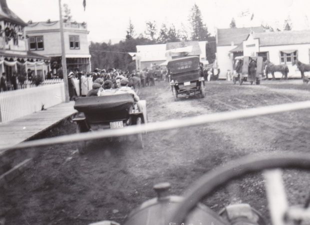 Une foule regarde un défilé du trottoir. Il y a des chevaux et des vieilles voitures dans le défilé. La rue est faite de terre et garnie de maisons et de petites entreprises.