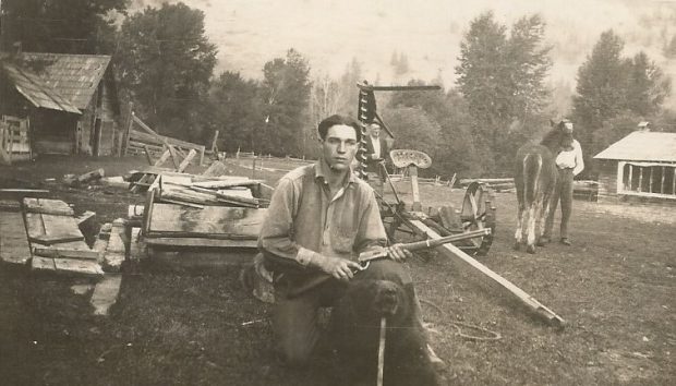 Un jeune homme tient un fusil dans la cour d’une ferme. Il a le genou droit sur le sol et il y a un ours noir derrière lui. L’ours a été abattu et sa tête est appuyée par un bâton. Il y a deux autres hommes dans l’arrière-plan. Un des hommes tient un cheval.