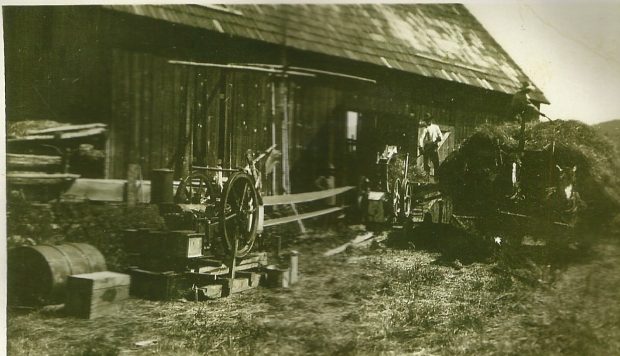 Deux chevaux sont attelés à un wagon plein de foin. Les chevaux sont debout à côté d’une étable. Deux hommes s’en servent une bande transporteuse et des fourches pour décharger le foin.