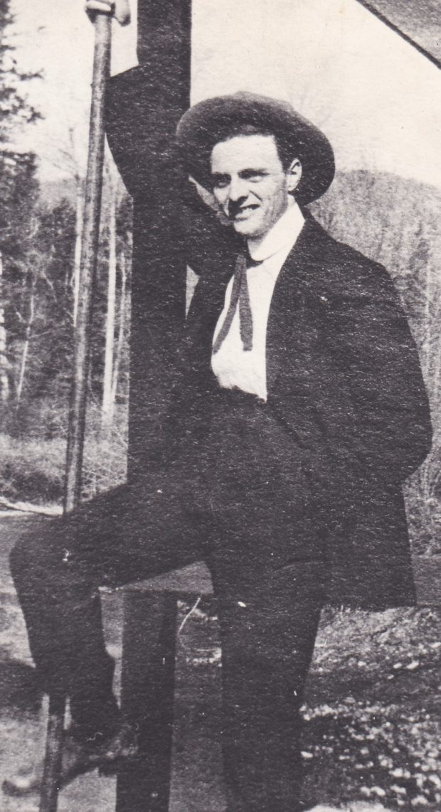 Un homme porte un costume noir et une cravate. Il pose devant la véranda d’une maison. Il porte aussi un chapeau. La maison semble être au milieu d’une région sauvage car s’est entouré par des arbres.