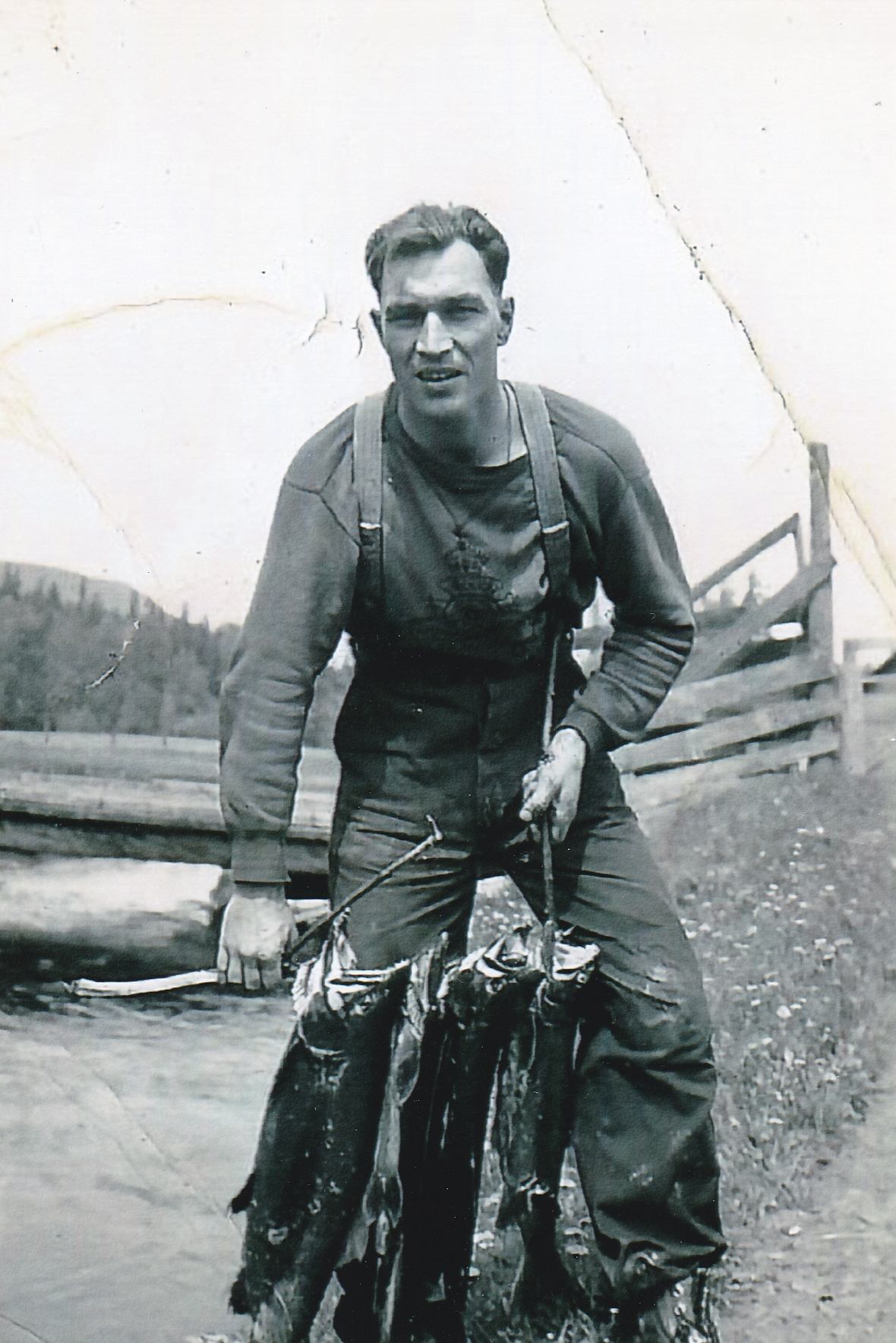 Un jeune homme tient un bâton avec trois saumons percés. L’homme est debout à côté d’une rivière. Il y a un pont avec des planches en bois dans l’arrière-plan.